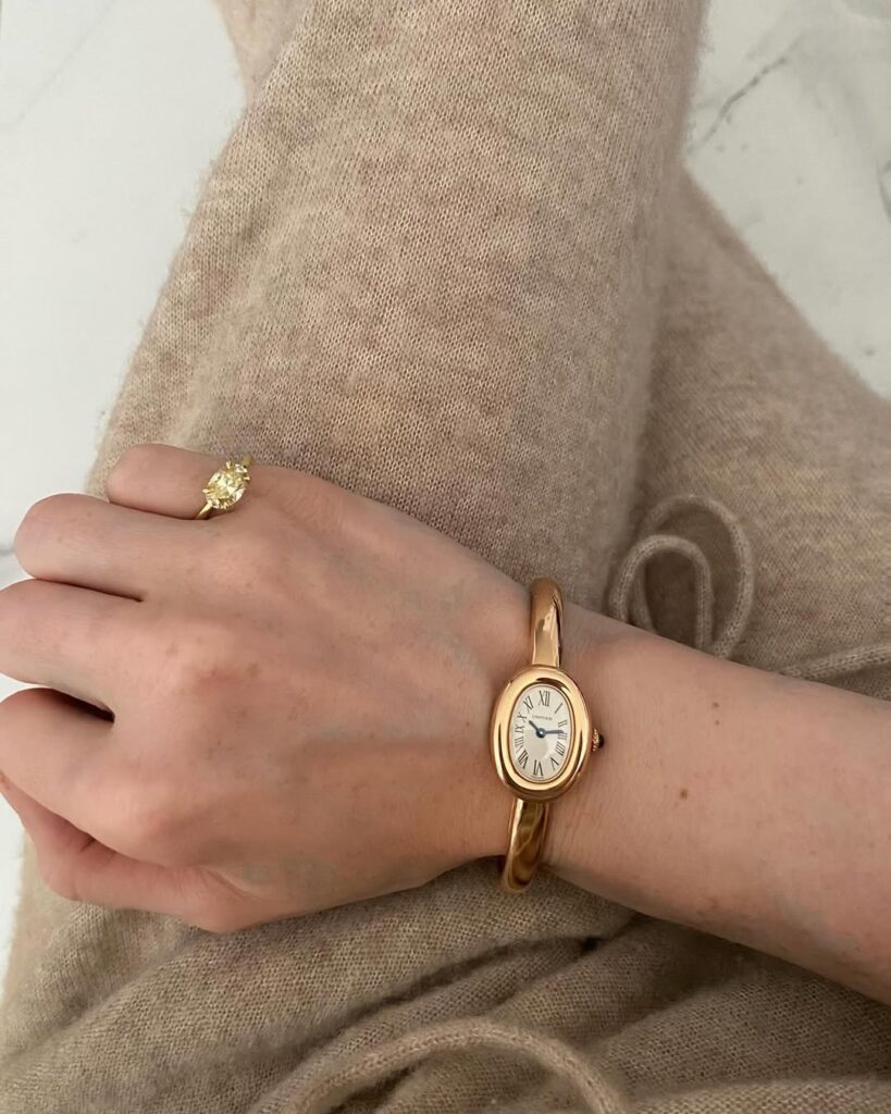 A close-up of a woman wearing a sleek gold wristwatch with a minimalist oval dial and Roman numerals, styled with a beige knit sweater and a simple gold ring. The soft neutral palette and polished finish create a warm, elegant quiet luxury aesthetic — perfect for daily sophistication.  